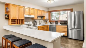 A modern kitchen with wooden cabinets, a stainless steel refrigerator, a microwave, and a large island with bar stools. - Top Floor and Spacious, One block to Everything! (Breckenridge)