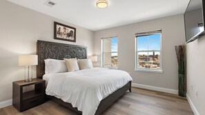 A bedroom with a large bed, two bedside tables with lamps, a decorative headboard, a framed picture, two windows with city views, and a tall plant. - City Views 1BR Free-Parking- Close to NYC (Newark)