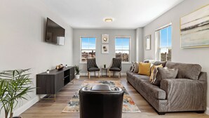 A modern living room with a grey sofa, two armchairs, a black ottoman, a dark wooden cabinet, and a flat-screen TV mounted on the wall. There are two windows with city views, a ceiling light, and a large abstract painting on the wall. - City Views 1BR Free-Parking- Close to NYC (Newark)