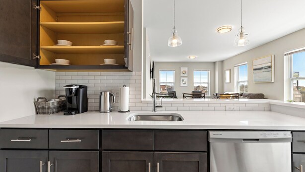 A modern kitchen with dark cabinets, a white countertop, a built-in oven, and a dishwasher. There are hanging pendant lights and a view of a dining area with a table and chairs. - City Views 1BR Free-Parking- Close to NYC (Newark)