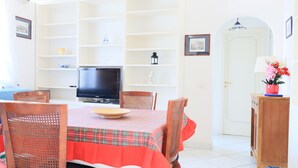 A dining area with a table set for four, surrounded by wicker chairs, a television on a stand, and shelves with various items. - Cataldo guest house Capri center (Marina Grande)