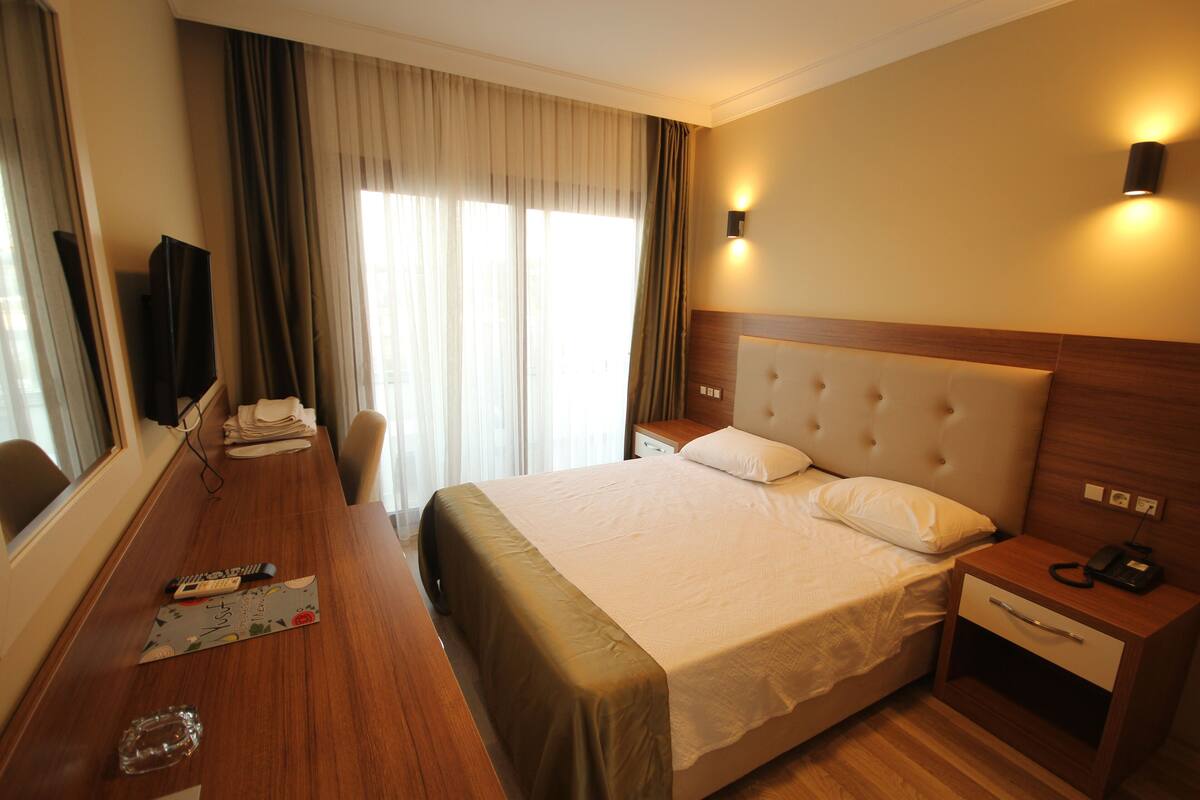 A hotel room with a large bed, a wooden desk, a chair, a bedside table with a lamp, and a window with curtains.