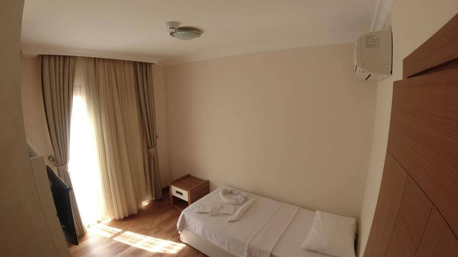 A small, well-lit room with a single bed, a wooden nightstand, a small table, and a window with curtains.