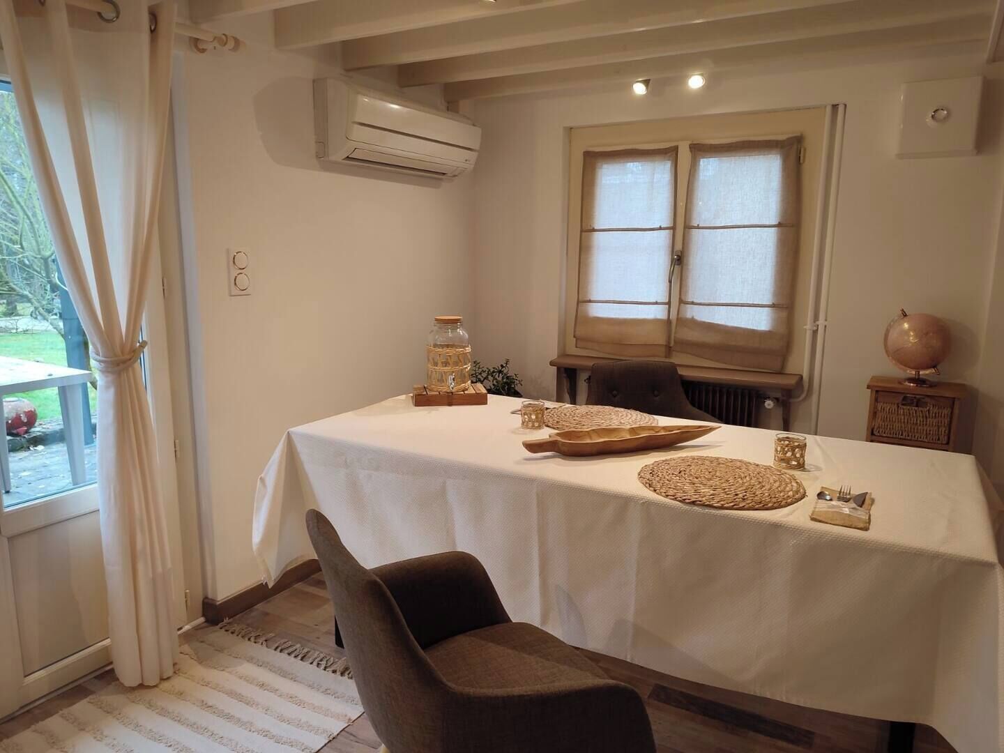 A dining area with a table set for two, featuring a white tablecloth, woven placemats, and a glass jar centerpiece.