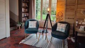 Een gezellige woonkamer met twee donkerblauwe fauteuils, een klein bijzettafeltje en een boekenkast. Een groot raam met gordijnen en uitzicht op groen is zichtbaar.