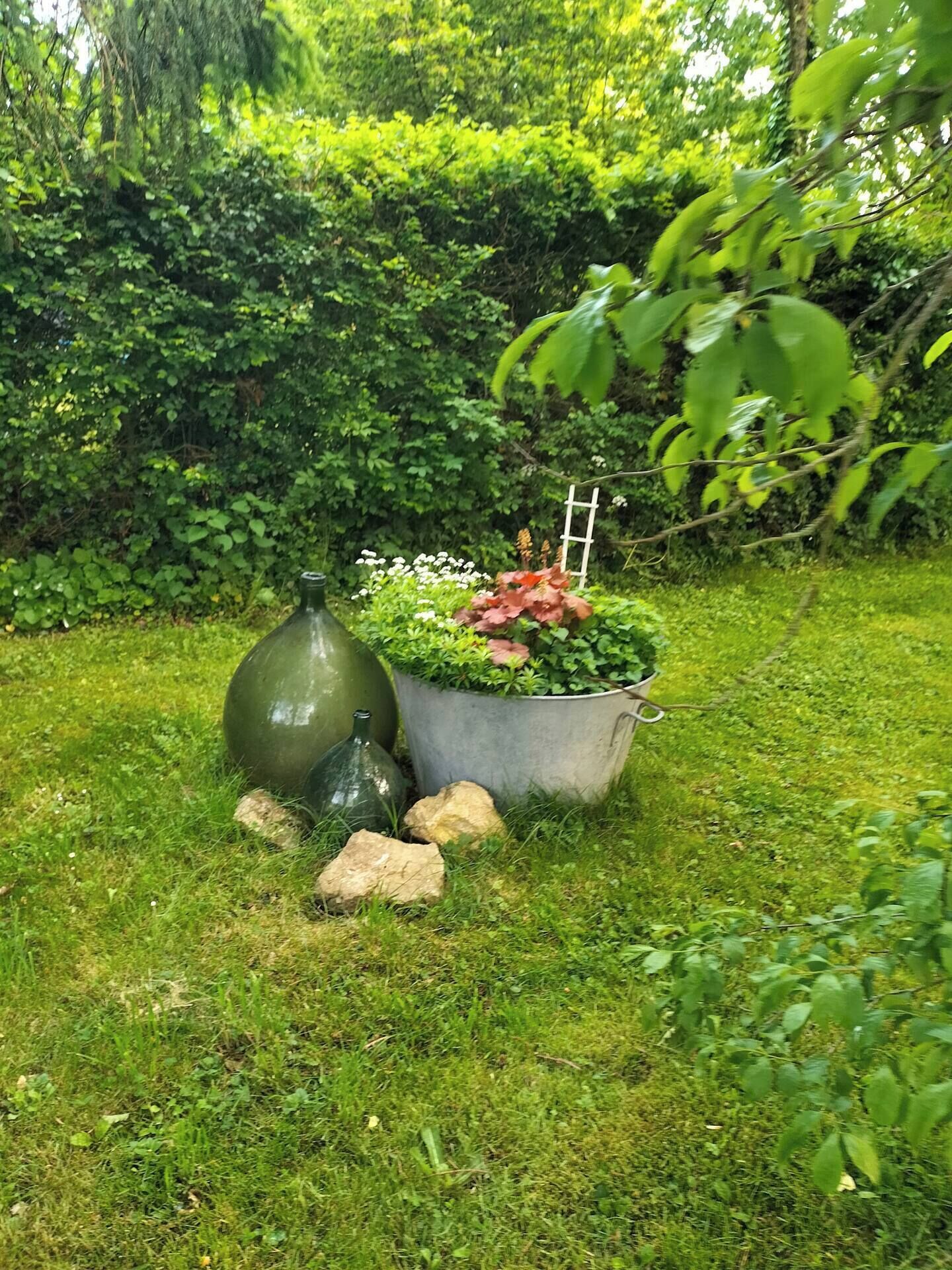 Un jardin agrémenté d’un grand vase vert, d’un bac en métal contenant des plantes et de rochers dispersés sur la pelouse.