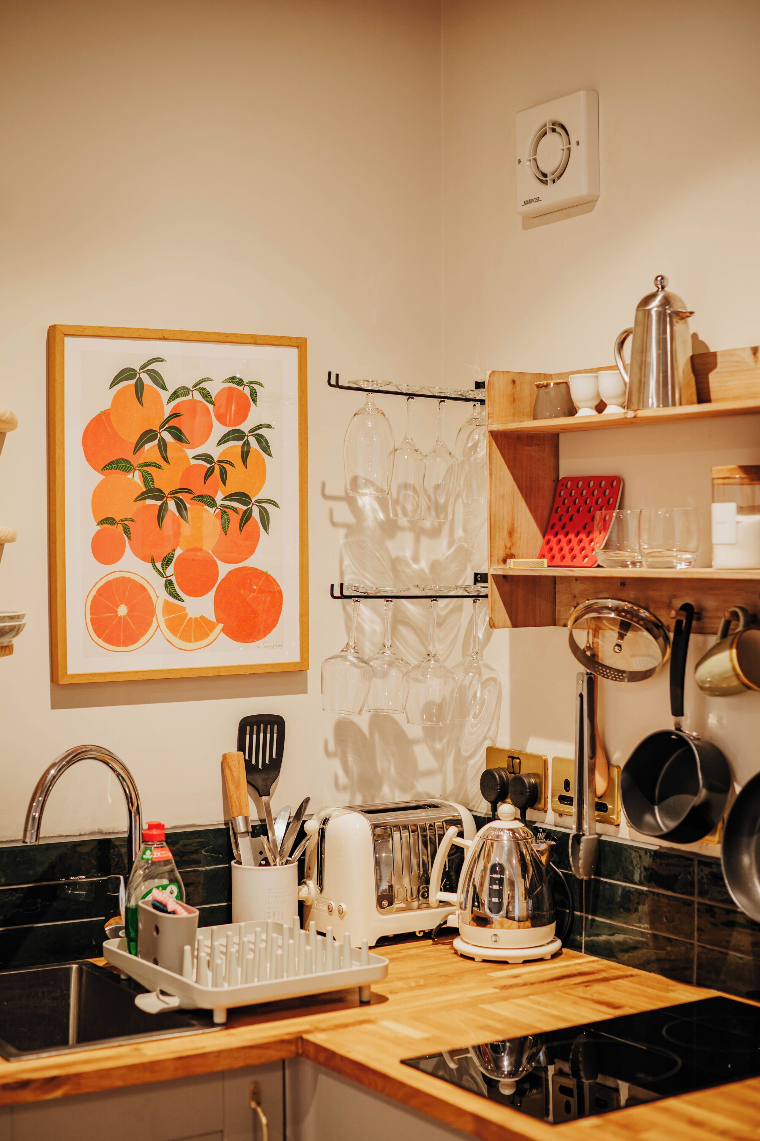 Une cuisine avec un plan de travail en bois, un évier, une cuisinière et divers ustensiles de cuisine. Un tableau encadré représentant des oranges est accroché au mur.