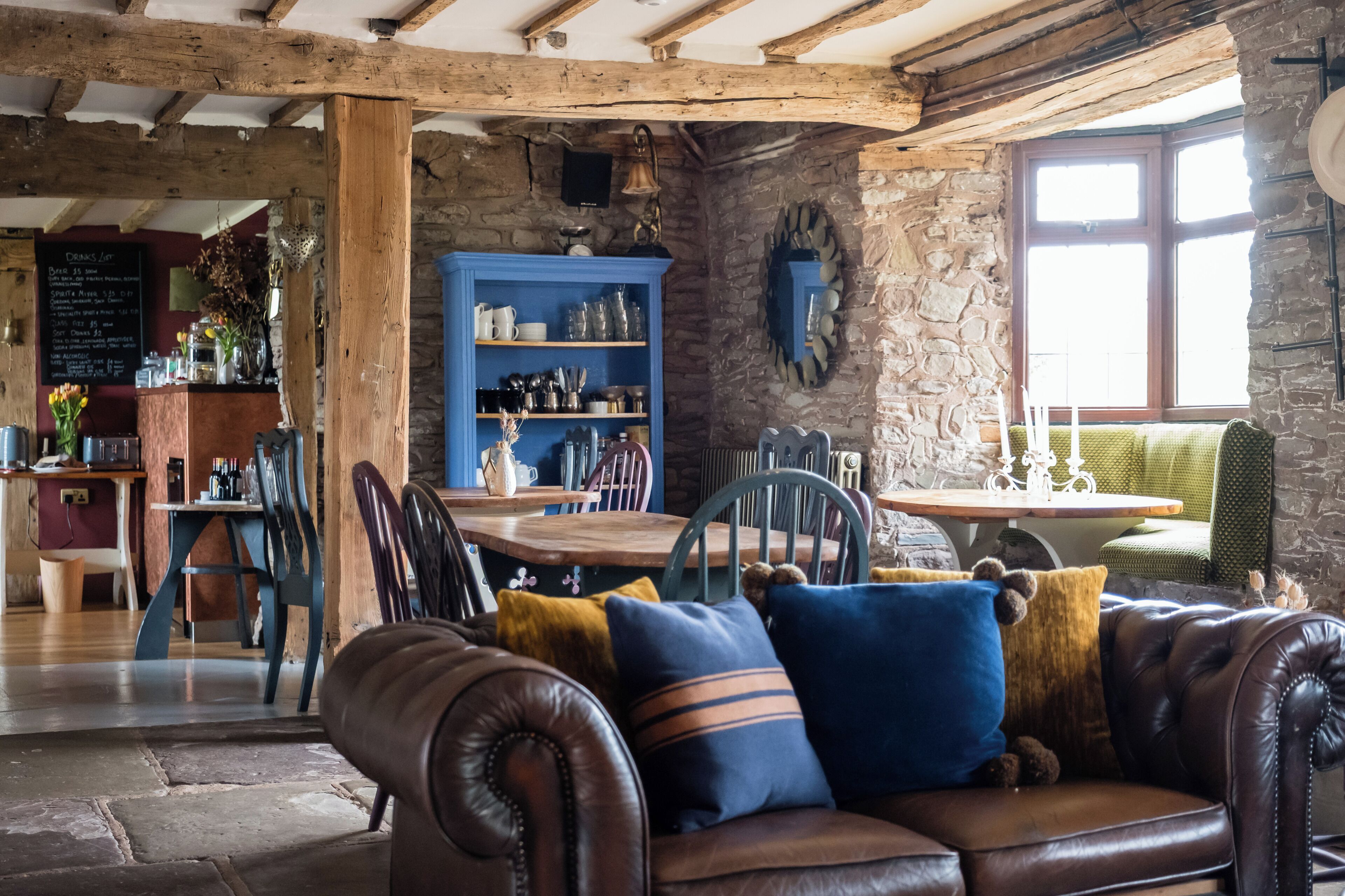 A rustic interior with exposed wooden beams, stone walls, a leather sofa with colorful cushions, and a stone fireplace.