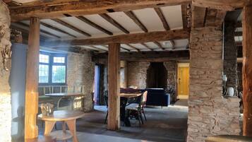 Ein rustikales Interieur mit freiliegenden Holzbalken, Steinwänden und einem Kamin aus Stein. Im Vordergrund steht ein runder Holztisch mit Stühlen und einer Bank.
