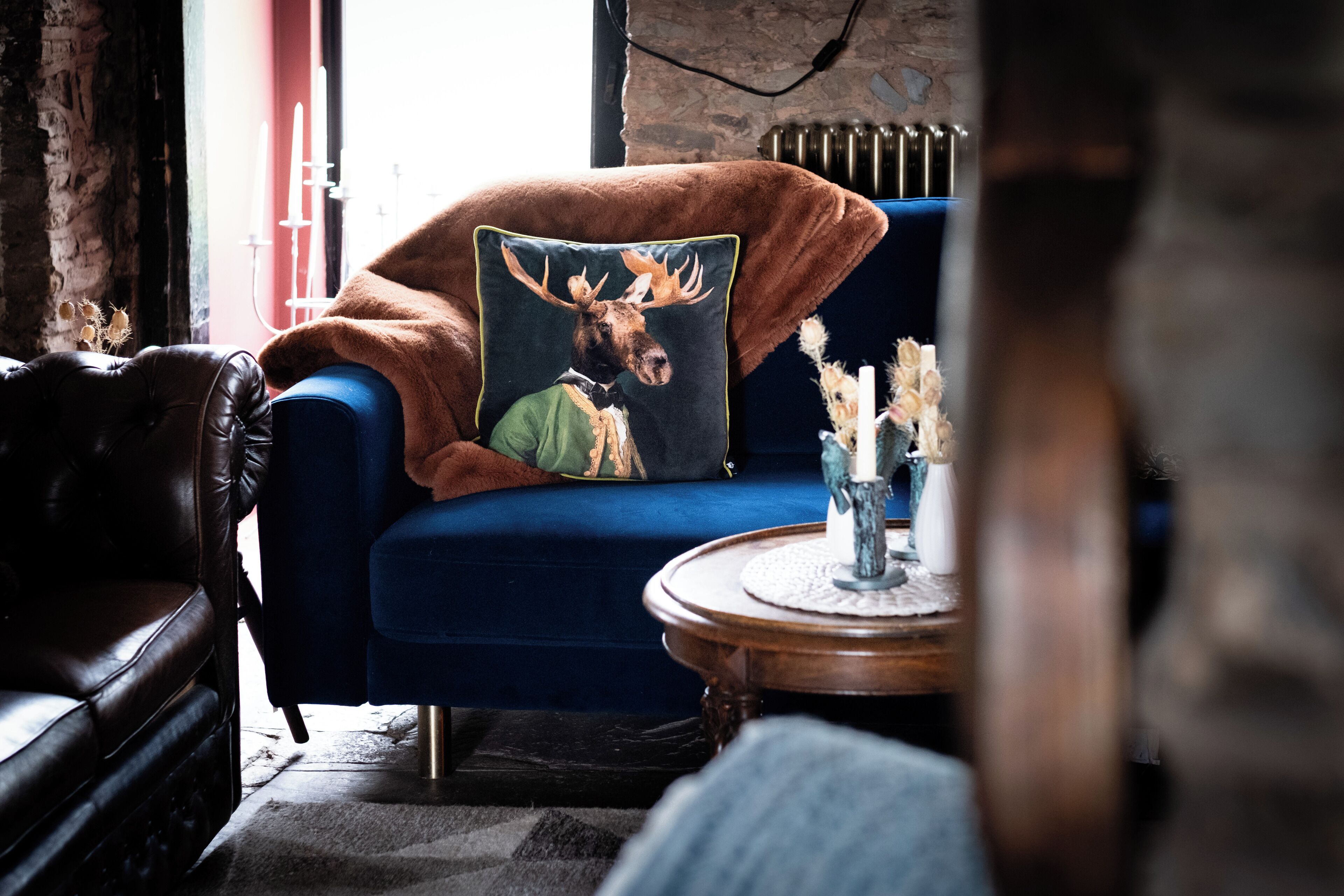 A cozy room with a blue armchair, a decorative pillow with a moose, and a small round wooden table with candles.