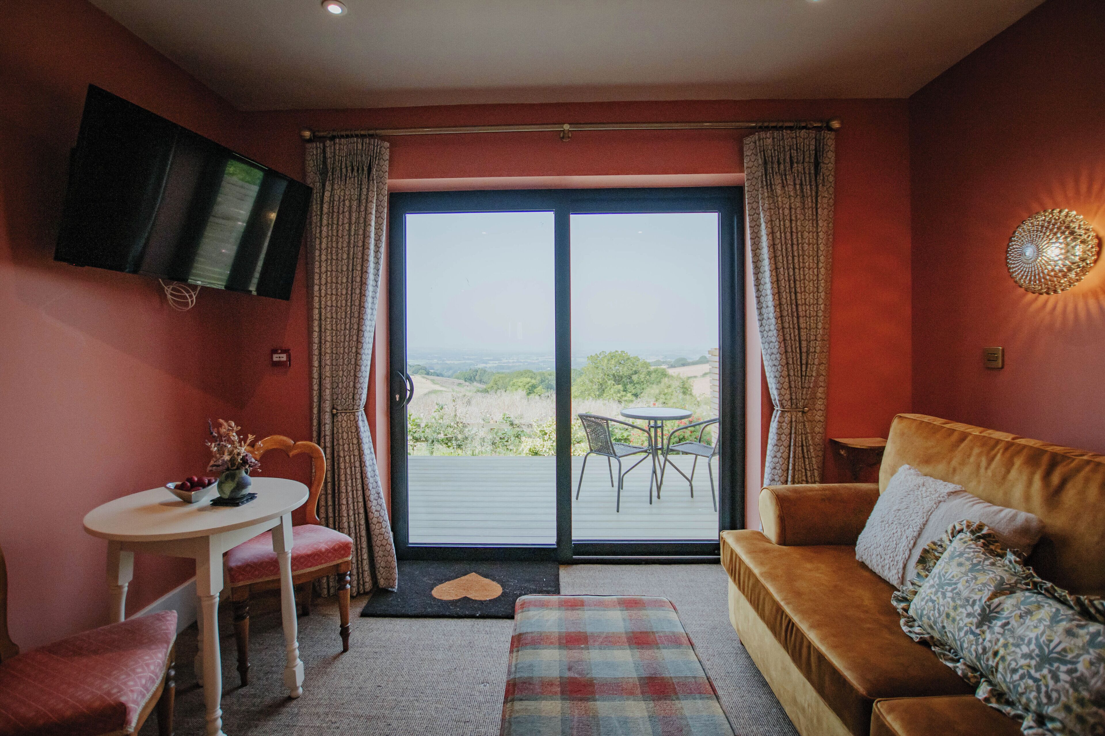 A cozy living room with a sofa, a small table with a bowl of fruit, a TV mounted on the wall, and a sliding glass door leading to an outdoor patio.