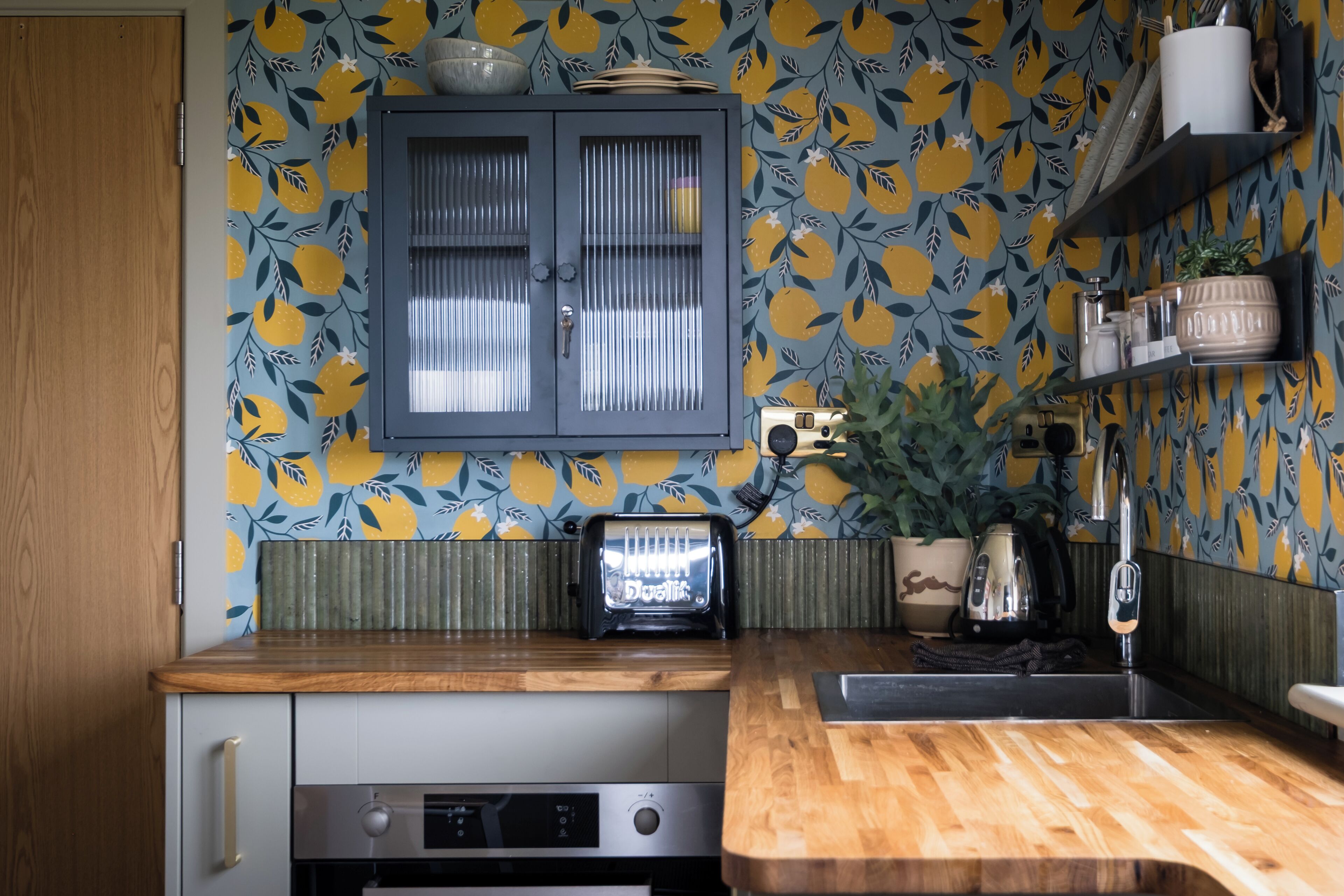 A kitchen with a wooden countertop, a built-in oven, a toaster, and a backsplash with a fruit pattern.