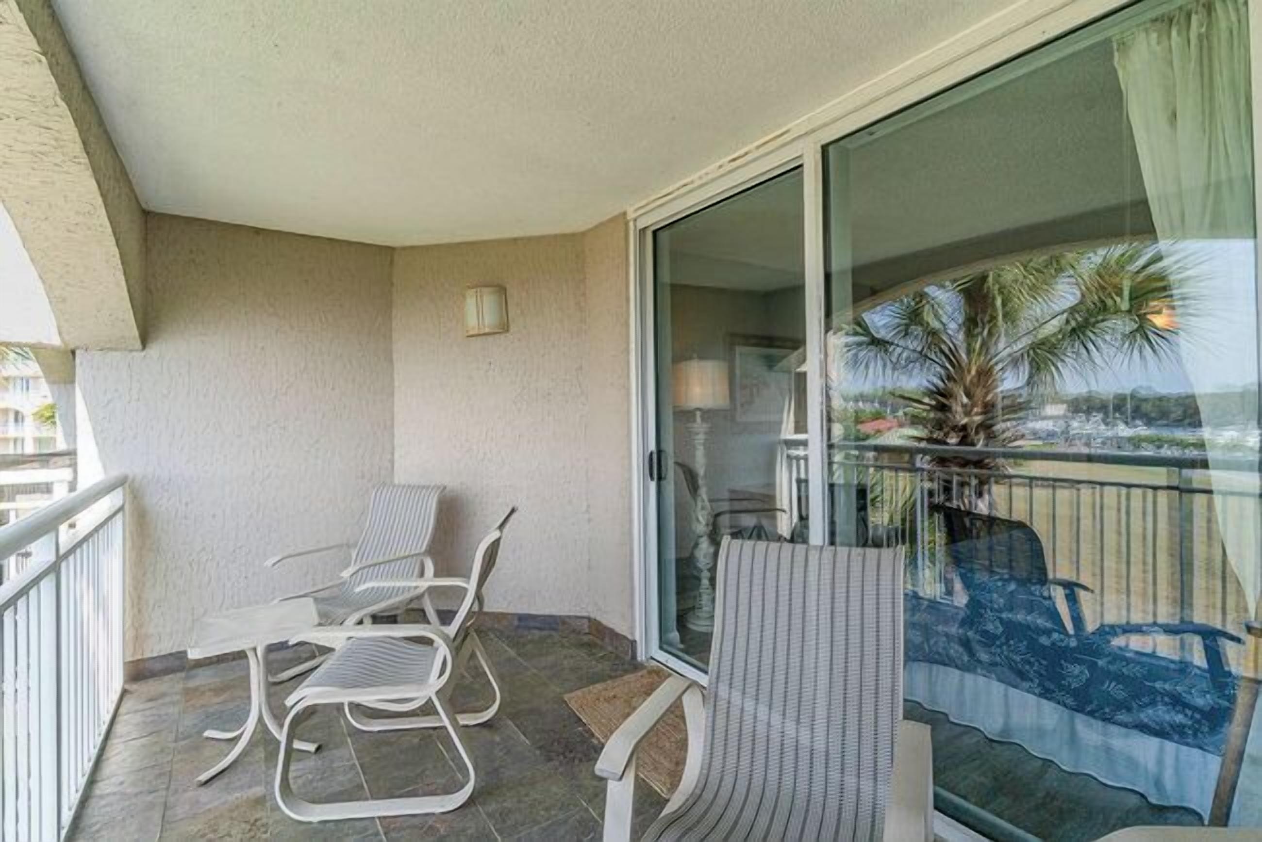 A balcony with white metal chairs and a table, a sliding glass door, and a view of palm trees and a beach.