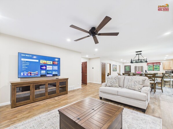A spacious living room with a flat-screen TV, wooden furniture, and a ceiling fan.