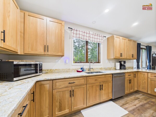 Una cocina moderna con armarios de madera, microondas integrado, lavaplatos de acero inoxidable y ventana con vistas a árboles.