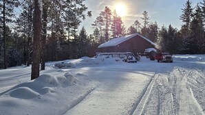 Una cabaña con techo nevado, rodeada de un bosque y un camino cubierto de nieve.