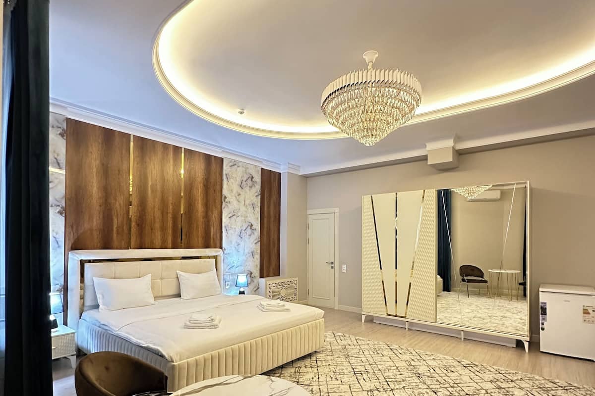 A modern hotel room with a large bed, a round table, two chairs, a chandelier, a large mirror, and a wardrobe.