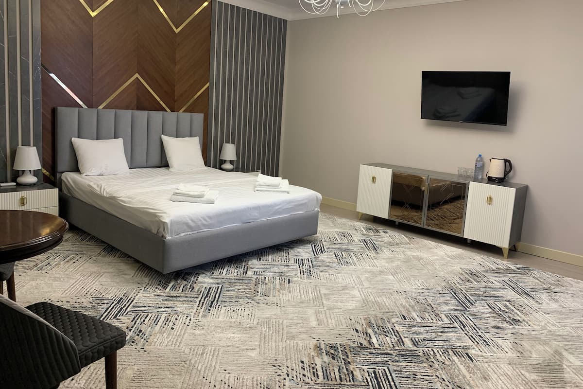A modern hotel room with a large bed, a bedside table with a lamp, a TV mounted on the wall, and a minibar.