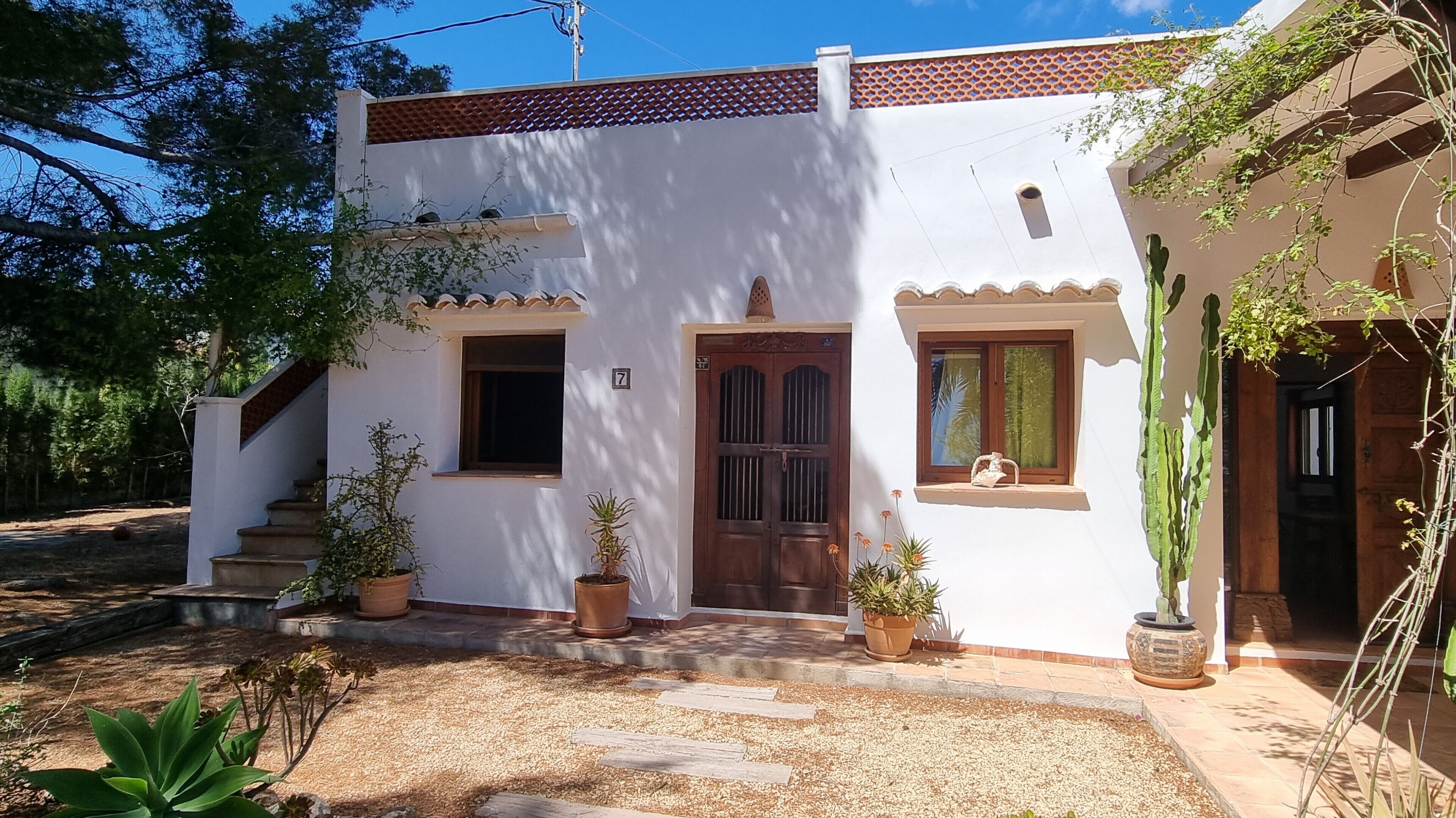 Un bâtiment blanc avec un toit recouvert de tuiles, une porte en bois et une fenêtre ornée d’un rideau. Des marches mènent à la porte, et plusieurs plantes en pot sont disposées autour de l’entrée.