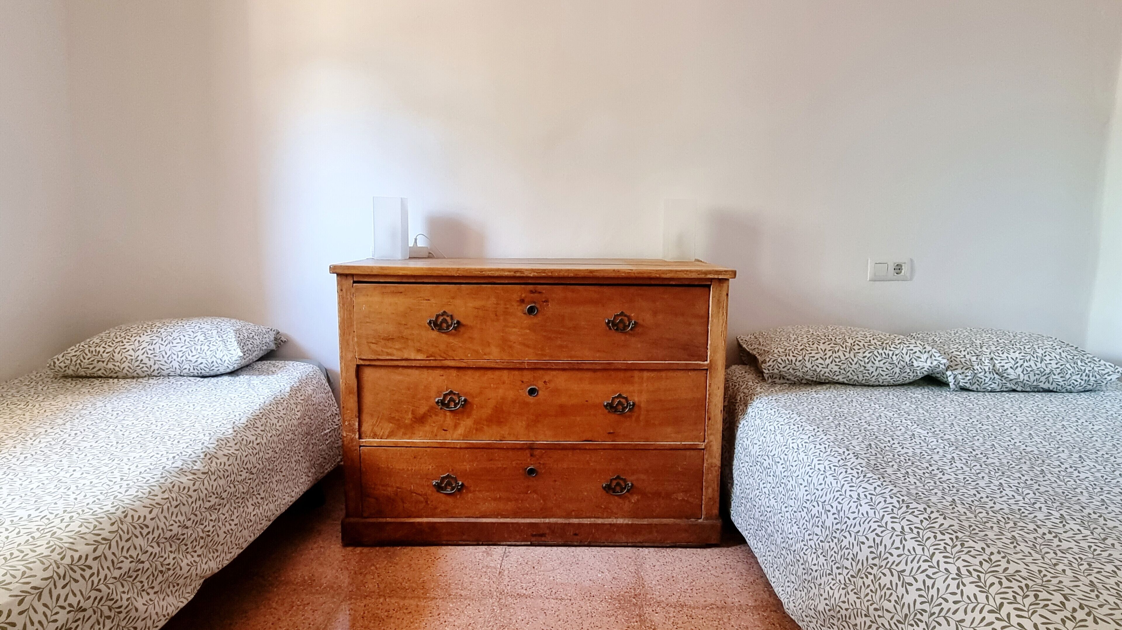 A room with two beds, each with patterned bedding, a wooden dresser with metal handles, and a white wall socket.
