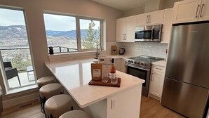 A modern kitchen with a central island, stainless steel appliances, and large windows offering a mountain view. - Bungalow in the Bluffs (Penticton)