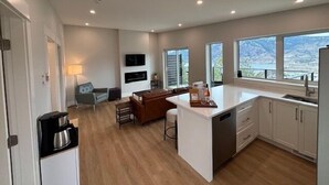 A modern kitchen with a central island, a dining area with a sofa and chairs, and large windows offering a view of the mountains. - Bungalow in the Bluffs (Penticton)