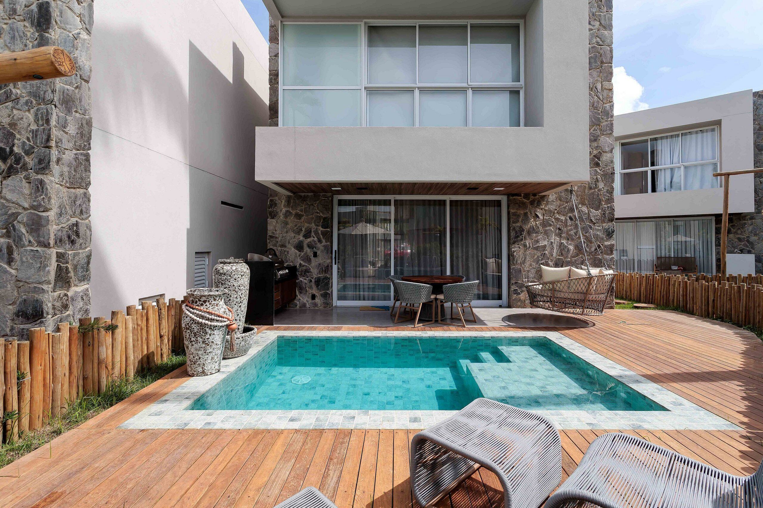 A modern outdoor pool area with a wooden deck, stone walls, and a seating area with wicker chairs.