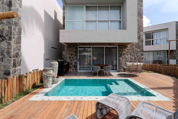 A modern outdoor pool area with a wooden deck, stone walls, and a seating area with wicker chairs. - Casa Kumari - By Living Milagres (Passo de Camaragibe)