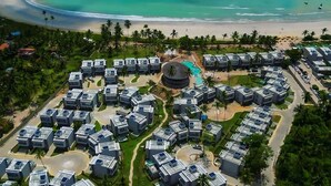 Luchtbeeld van een resort met meerdere moderne gebouwen, een centraal gelegen circulair zwembad en een strand met helder turquoise water.
