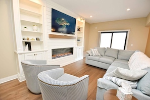 Un salon avec une cheminée, un canapé gris, deux fauteuils en osier et un téléviseur fixé au mur.