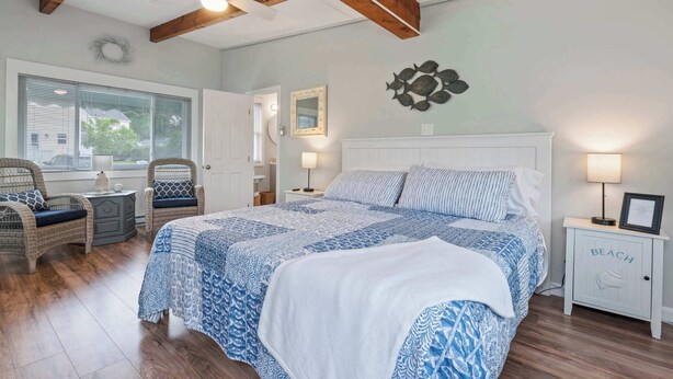 A bedroom with a large bed, two bedside tables, wicker chairs, a ceiling fan, and a view through a window.