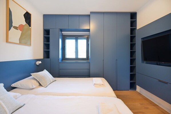 A modern bedroom with two beds, a built-in TV, and a window.