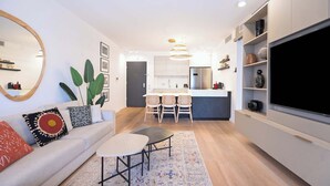 Una sala de estar moderna con sofá, mesa de centro y televisor. Se ve una cocina con zona de comedor al fondo.