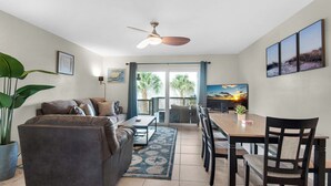 Un salon comprenant un canapé, une table basse et une table à manger avec des chaises. On y trouve également un ventilateur de plafond, une grande fenêtre offrant une belle vue et une plante en pot.