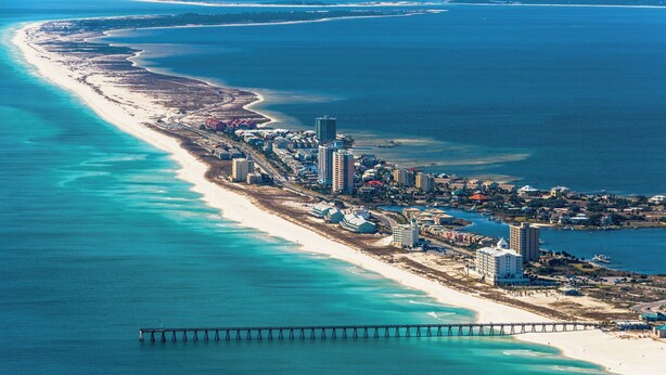 Une vue aérienne d’une ville côtière, avec une longue plage de sable, une jetée qui s’avance dans l’eau et plusieurs immeubles de grande hauteur le long de la côte.