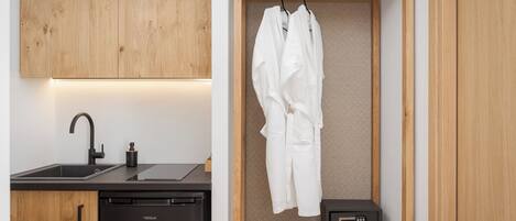 A compact kitchen with a dishwasher, a black sink, and a wooden cabinet. Adjacent to the kitchen is a small wooden shelf with a white robe and a black radio.