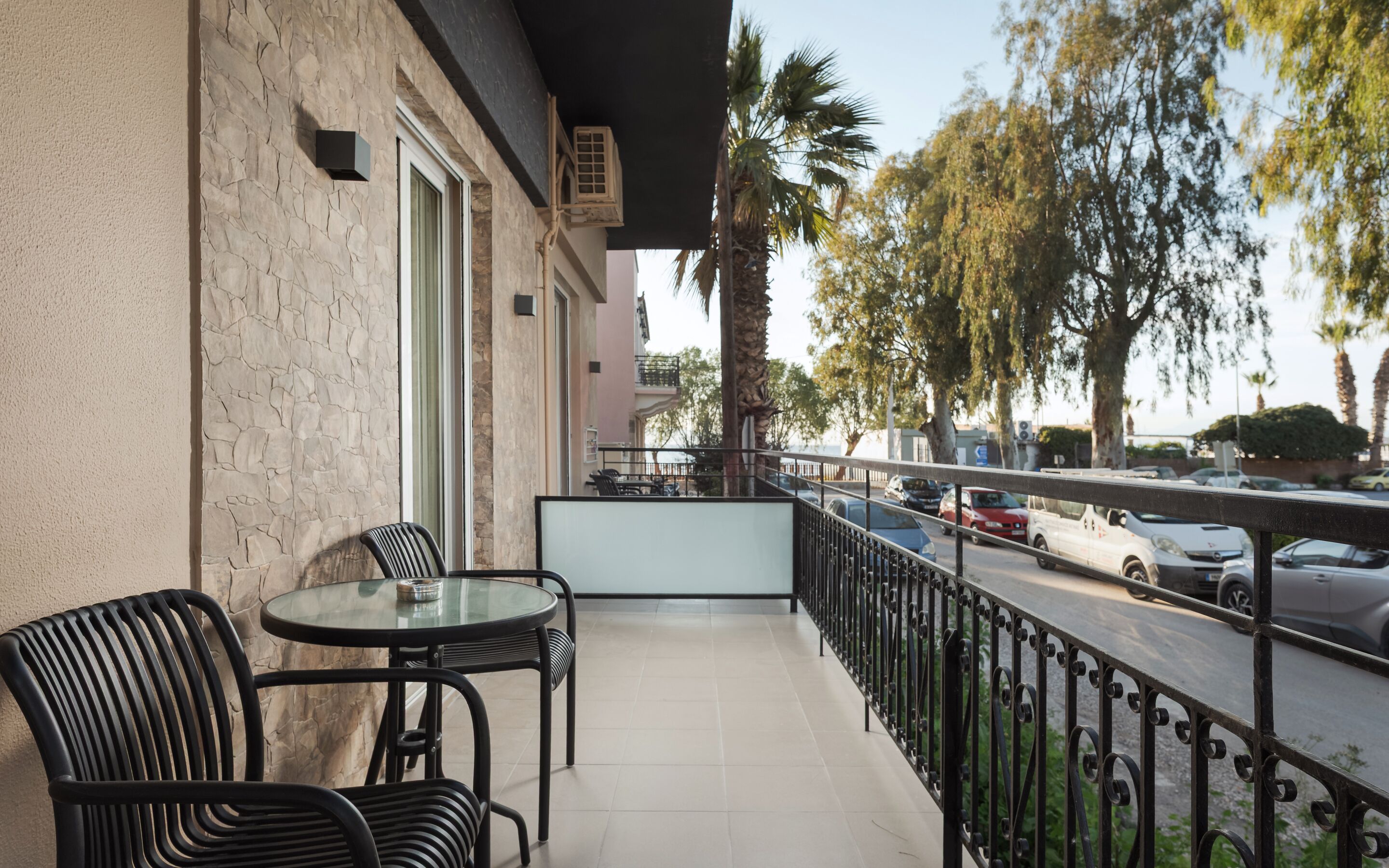 A balcony with a table and chairs, a view of parked cars, and a street with palm trees.