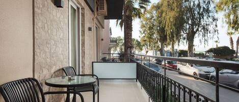 A balcony with a table and chairs, a view of parked cars, and a street with palm trees.