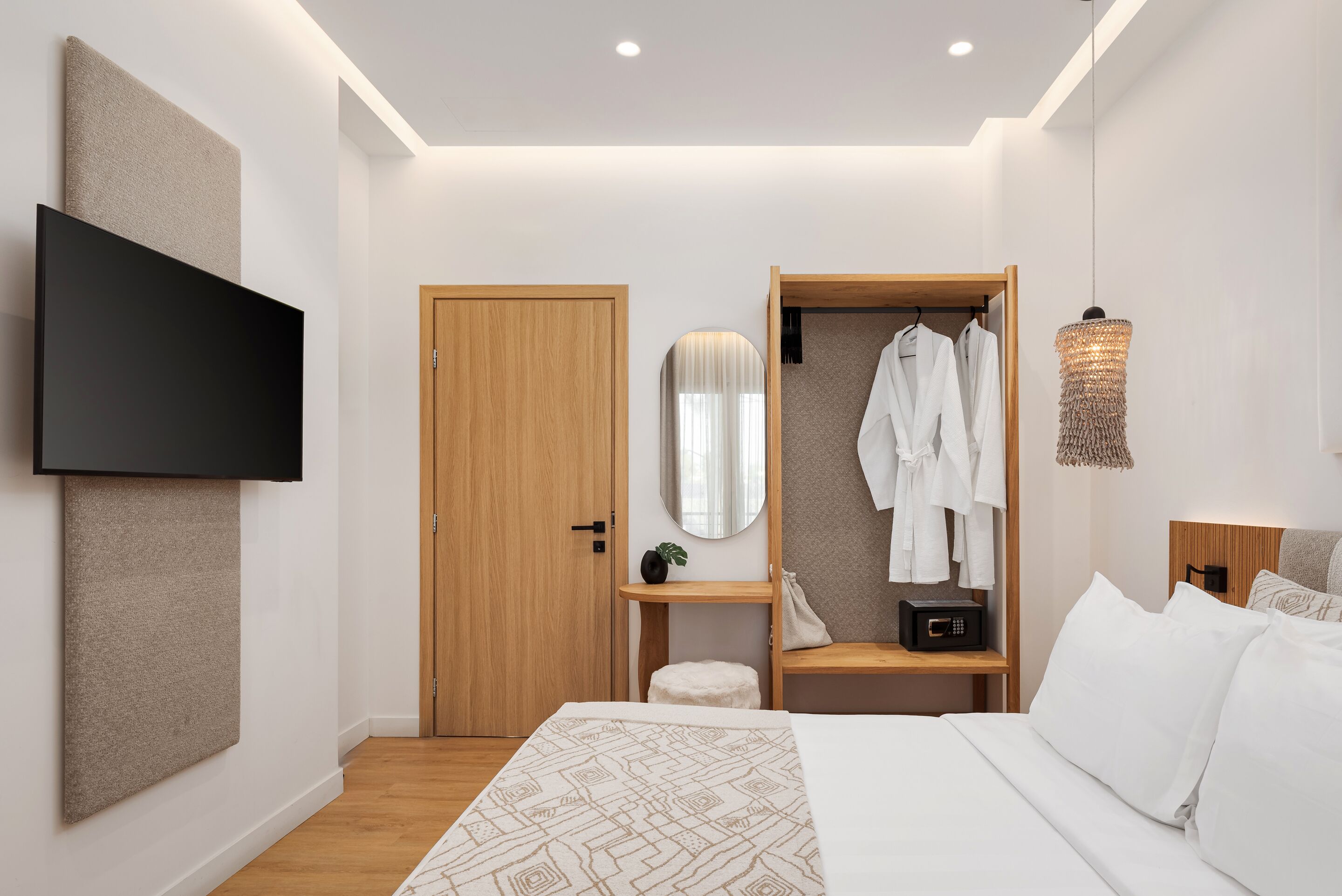 A modern hotel room with a bed, a TV mounted on the wall, a wooden wardrobe, a bedside table with a lamp, and a mirror.
