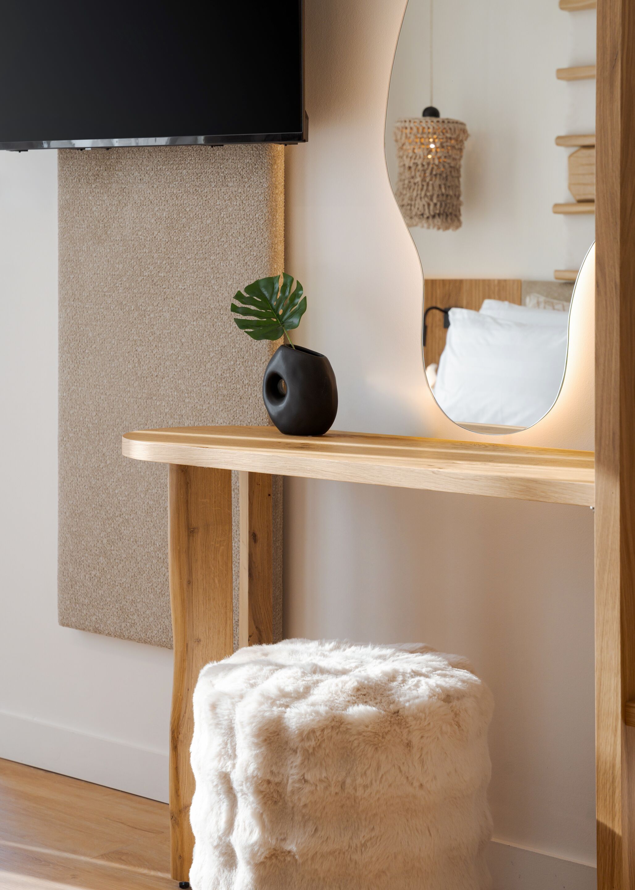 Una camera moderna con pavimento in legno, un pouf bianco e soffice, un vaso nero con una pianta, una mensola in legno, una TV a parete e uno specchio che riflette un letto e una lampada a sospensione.