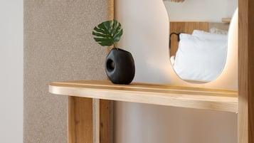 Una camera moderna con pavimento in legno, un pouf bianco e soffice, un vaso nero con una pianta, una mensola in legno, una TV a parete e uno specchio che riflette un letto e una lampada a sospensione.