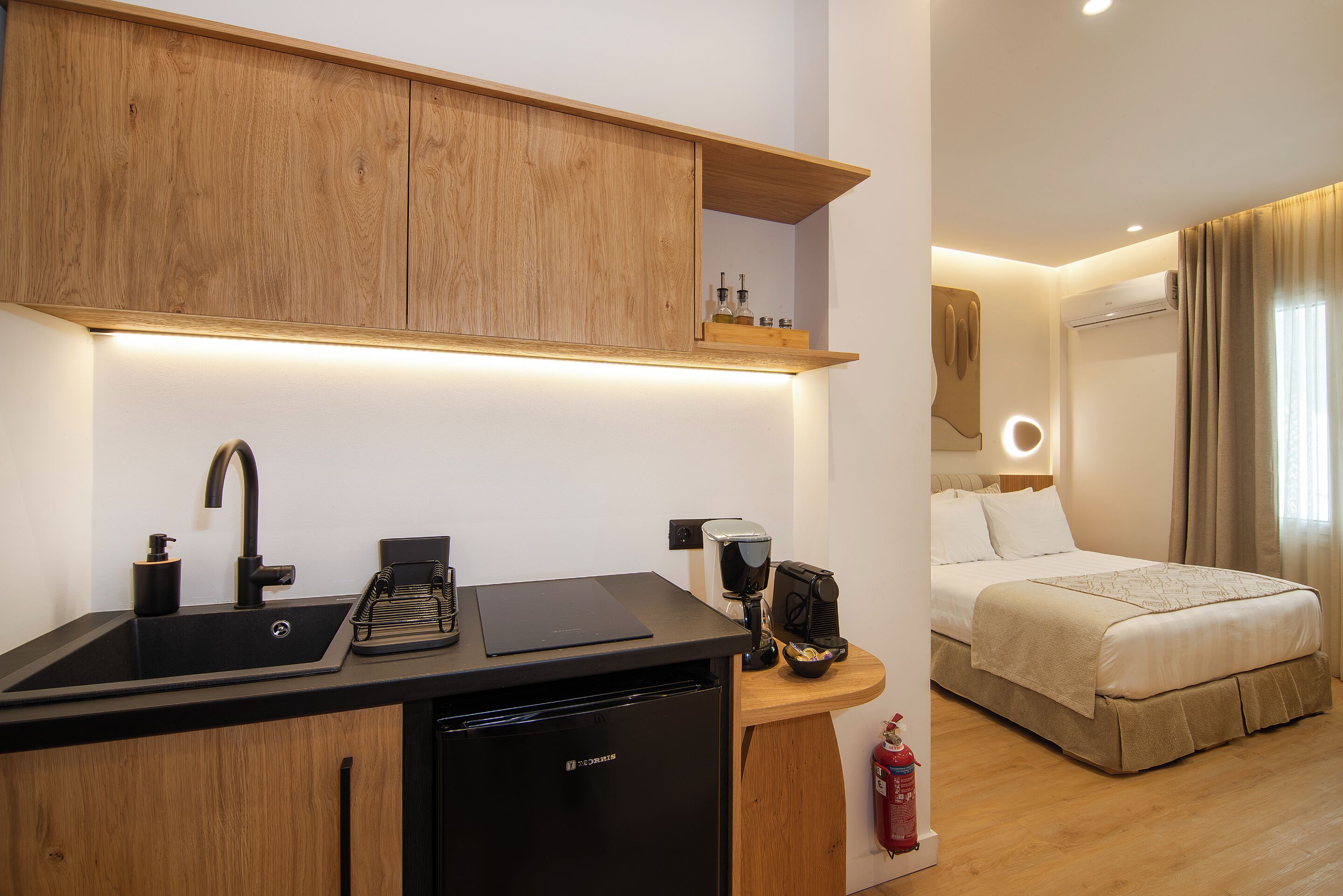 A compact hotel room with a kitchenette, a bed, and a bedside table.