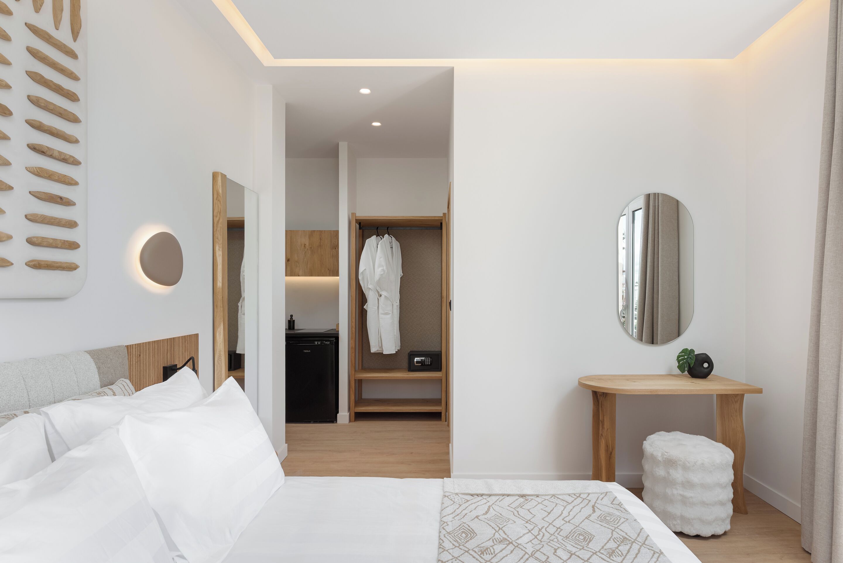 Une chambre d’hôtel moderne avec un lit soigneusement fait, une table de chevet en bois avec un miroir, un petit tabouret et un dressing où est suspendue une robe.