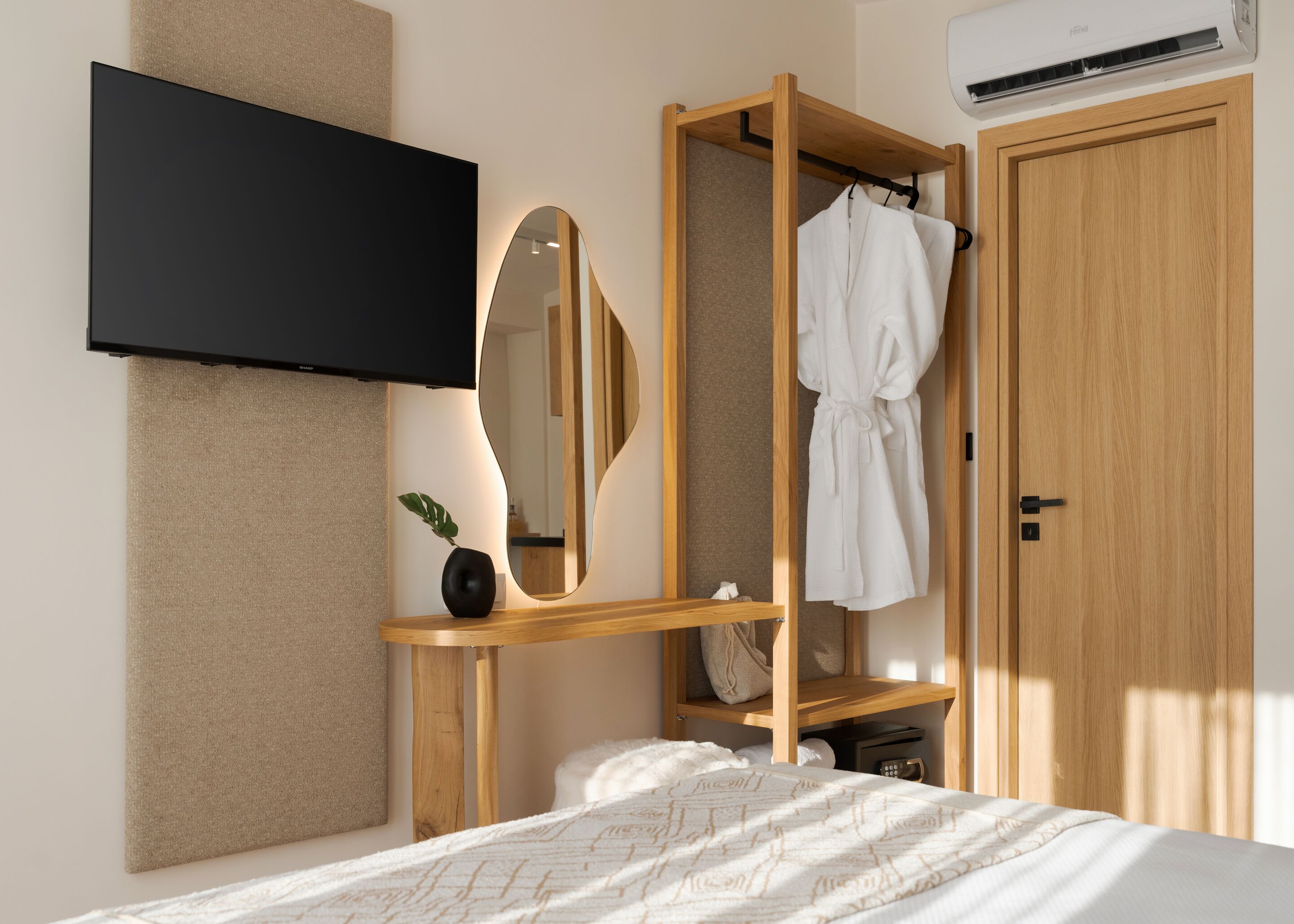 A modern bedroom with a bed, a wooden bedside table, a mirror, a robe hanging on a wooden rack, and a TV mounted on the wall.