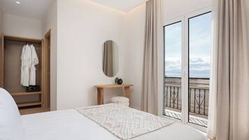 A modern hotel room with a large bed, a wooden wardrobe, a small table with a mirror, and a balcony with a view of the sea.