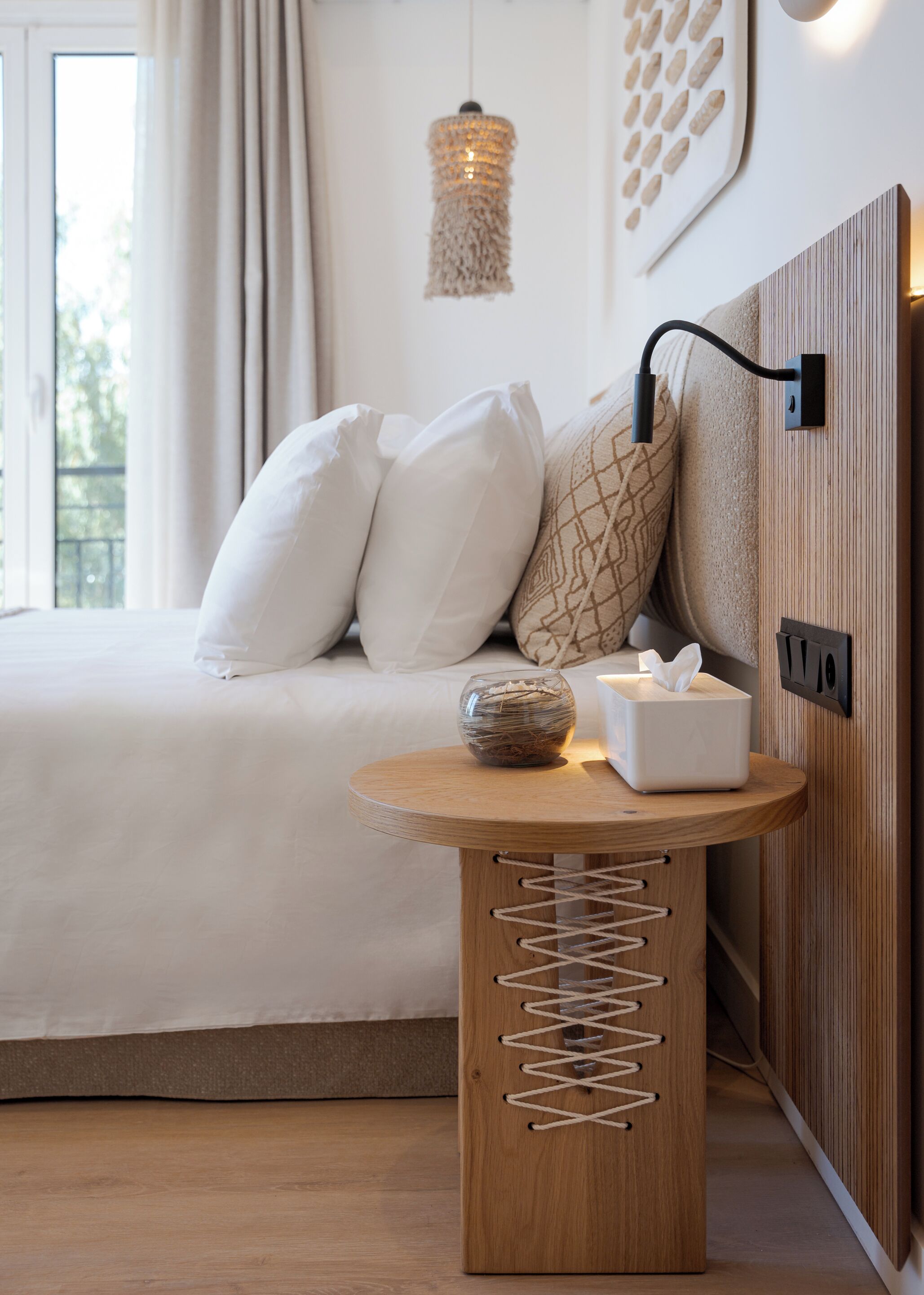 Un dormitorio ordenado con cama, almohadas, una mesita de noche con un dispensador de pañuelos, un cuenco de vidrio decorativo y una lámpara de pared.