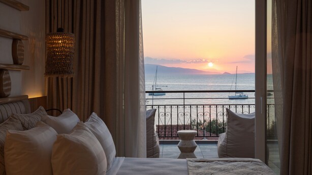 A hotel room with a bed, pillows, a hanging light fixture, and a view of the sea and boats at sunset. - Aegina Dream Suites (Aegina)
