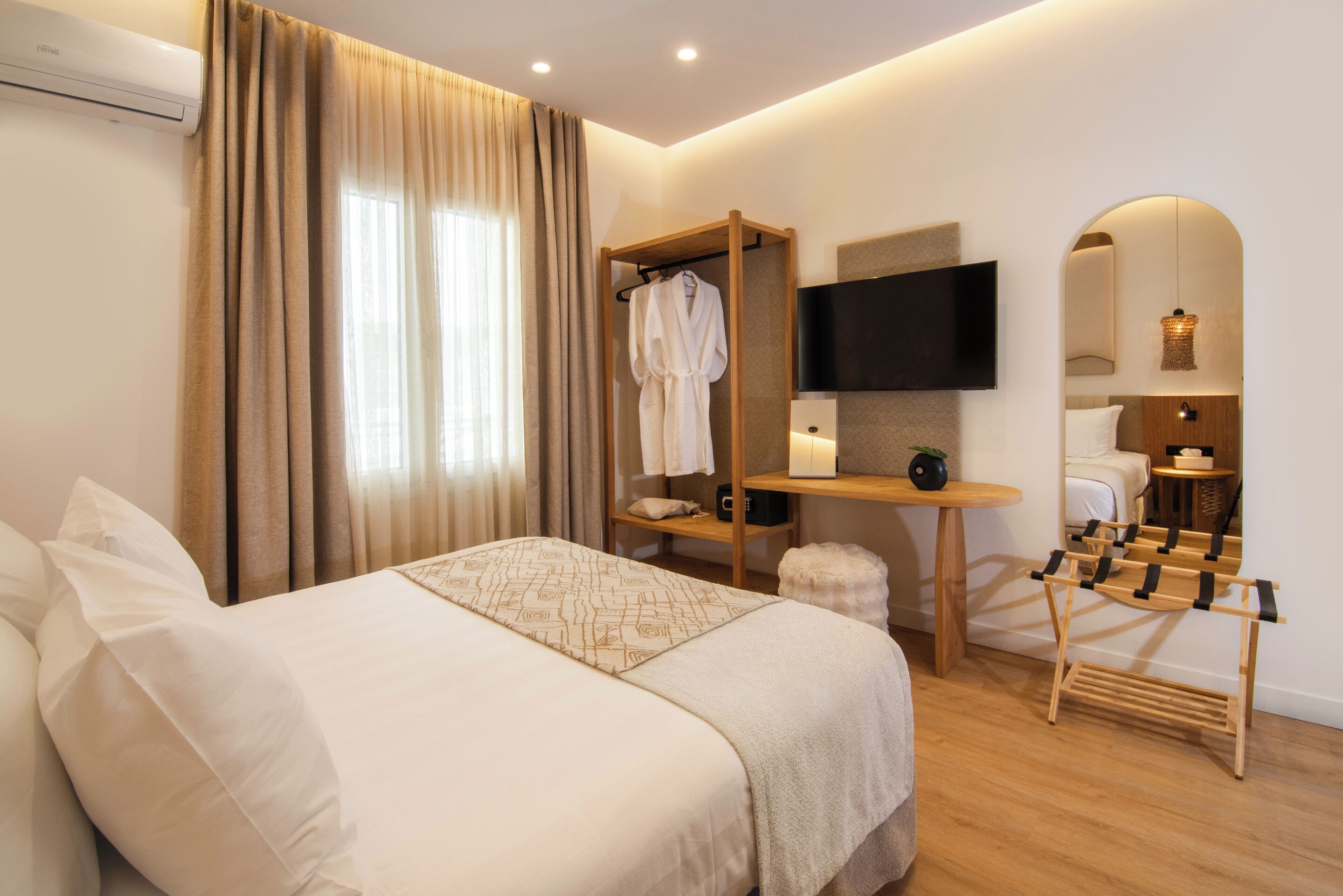 A hotel room with a large bed, a TV mounted on the wall, a wooden drying rack, and a small table with a lamp.