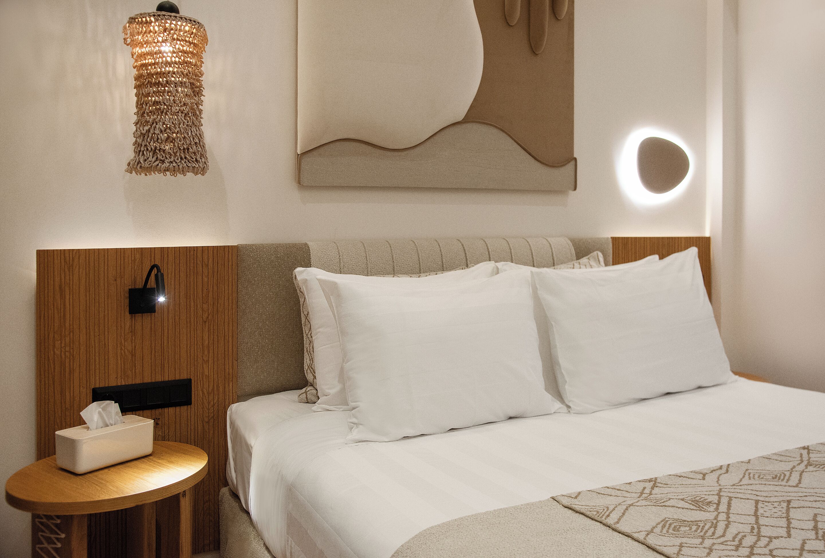 A neatly made bed with white linens, a bedside table with a tissue box, a hanging light fixture, and a wall-mounted light.