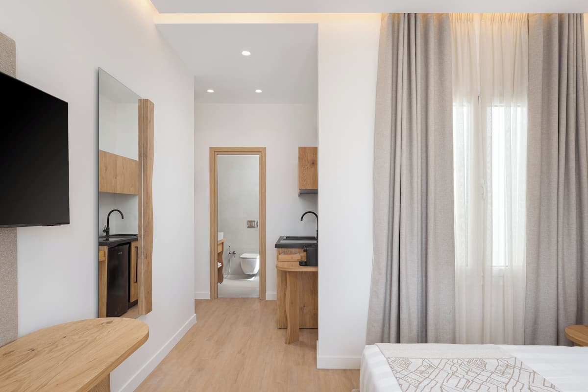 Une chambre d’hôtel moderne comprenant un lit, un banc en bois, une télévision, une salle de bain avec des toilettes et un lavabo, et une vue par une porte.